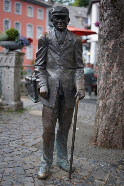 Denkmal für einen einfachen Mann
Maaßens Päulche, wie Paul Maaßen genannt wurde, verdiente sich sein tägliches Geld als reisender Kaufmann, indem er Kurzwaren aus seinem Korb heraus an den Haustüren anbot und verkaufte. Er soll dabei nicht nur in Monschau unterwegs gewesen sein. Sein Einzugsgebiet soll sich bis Aachen erstreckt haben. Doch hier in Monschau war er sehr beliebt. Denn Päulche hatte immer was zu erzählen und ständig einen netten Spruch parat. Das ein oder andere Mal haben ihm deshalb die Monschauer Frauen etwas abgekauft, auch wenn sie gerade nichts benötigten. Maaßens Päulche lebte von 1856 bis 1940 und wurde damit 84 Jahre alt. Die Menschen erzählten sich, dass Päulche sich anscheinend die letzten 20 Jahre kaum äußerlich merkbar verändert hatte. Das lag sicherlich an seinem Humor, seinem Hut und der frischen Luft, an der er ständig war. Oder war es gar der 29. Februar, an dem er geboren sein soll. Somit konnte er ja nur jedes 4. Jahr älter werden!
Gestiftet wurde die Skulptur durch  den Kunstkreis Nordeifel!
